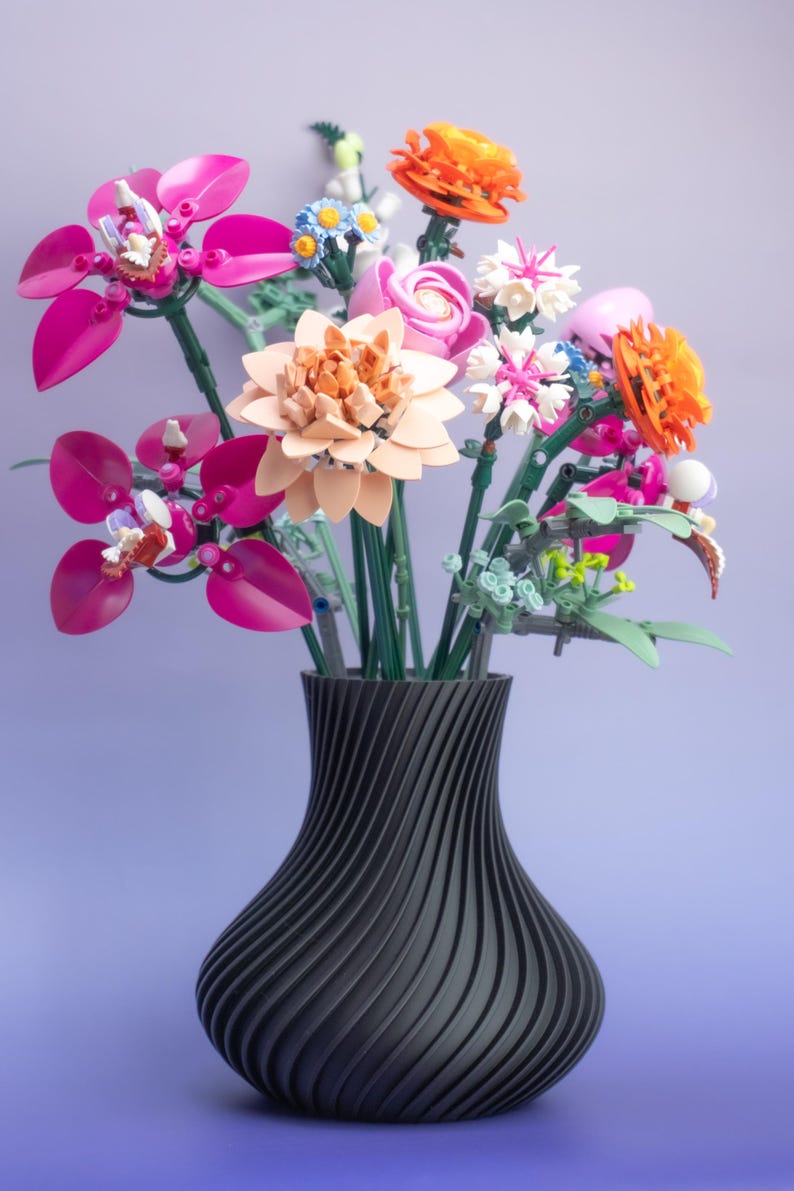 May include: A black vase with a swirling design holds a vibrant bouquet of colorful flowers. The flowers include pink, orange, and white blooms, with green stems. The arrangement is set against a gradient purple background.