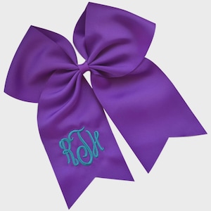 May include: A large, purple hair bow with a grosgrain texture. The bow features a teal monogram of the letters RTH embroidered on one of the tails. The bow is a classic accessory for cheerleading or everyday wear.
