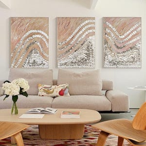 Peut inclure: Un salon avec trois peintures abstraites aux tons roses et argentés au-dessus d'un canapé beige. Deux chaises en bois et une table basse reposent sur un tapis à motifs. Un vase de fleurs blanches ajoute une touche d'élégance.