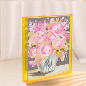 Könnte beinhalten: Ein quadratischer Acrylblock mit einem impressionistischen Gemälde eines Blumenstraußes aus rosa und weißen Blüten in einer klaren Vase. Das Kunstwerk ist vor einem dunkelgrauen Hintergrund platziert und von einem leuchtend gelben Acrylrahmen umgeben.