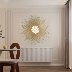 Könnte beinhalten: Eine goldene Sonnenstrahlen-Wandleuchte mit einer weißen Glaskugel als Leuchtmittel. Die Leuchte ist an einer weißen Wand mit einem weißen Heizkörper darunter montiert.