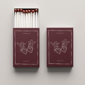 Pu&ograve; includere: Due scatole di fiammiferi bordeaux con un design decorativo. Il design presenta due cherubini che tengono un cuore, con il testo "October 19, 2010" e "Mona & John". Le teste dei fiammiferi sono marroni.