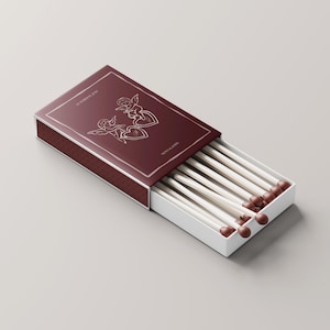 Pu&ograve; includere: Una scatola di fiammiferi bordeaux con interno bianco, parzialmente aperta per rivelare fiammiferi bianchi con punte marroni. Il coperchio della scatola presenta un contorno argentato e un disegno di due cherubini e cuori. Testo sulla scatola: "OCTOBER 2022" e "NOUVELLE LUNE".