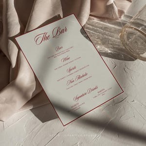Pu&ograve; includere: Un menu bianco con bordo rosso intitolato "The Bar" elenca diverse bevande alcoliche e analcoliche, tra cui birra, vino, liquori e cocktail firmati come Negroni e Martini.