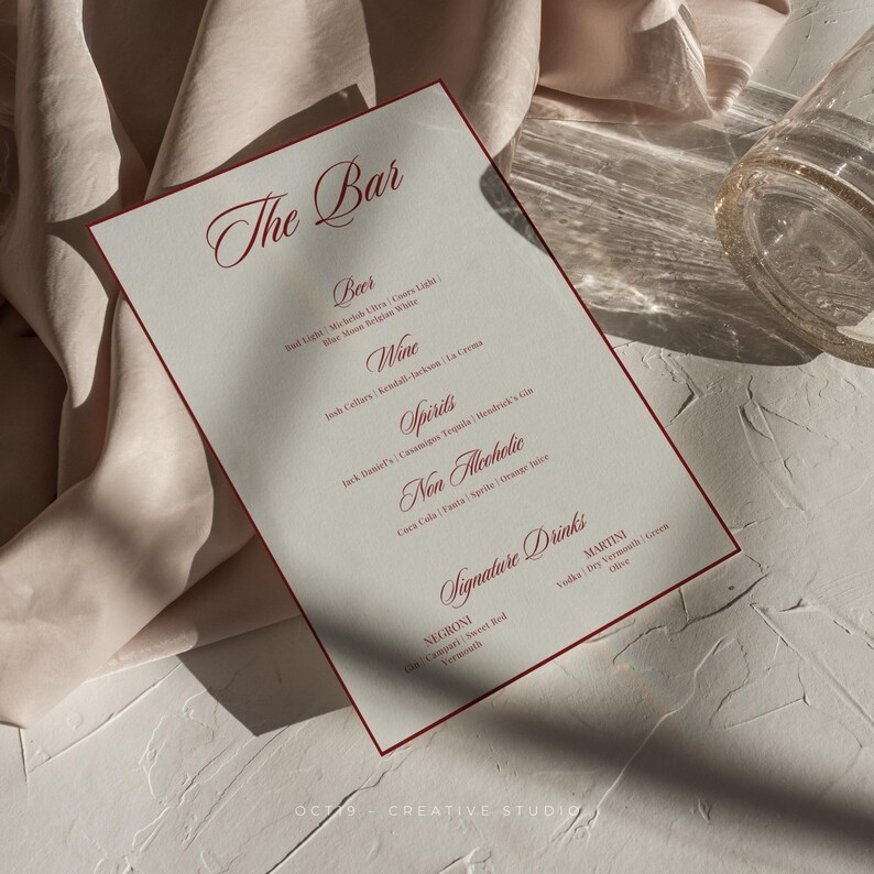 Pu&ograve; includere: Un menu bianco con bordo rosso intitolato "The Bar" elenca diverse bevande alcoliche e analcoliche, tra cui birra, vino, liquori e cocktail firmati come Negroni e Martini.