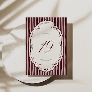 May include: A burgundy and cream table number card with vertical striped background. The card features an ornate cream-colored frame with the number 19 in burgundy. The text "TABLE NO." and "LOUISE & JAMES" are also visible.