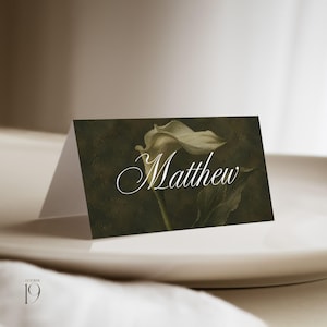 May include: A folded rectangular place card featuring a dark, moody green background with a white calla lily illustration. The name Matthew is printed in a white, elegant script font across the centre of the card, which stands upright.