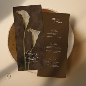 May include: A dark brown wedding menu card featuring two white calla lilies, the names Chiara & Michael, and the date October 19, 2028. The text lists courses including Smoked Salmon Blinis, Miso Marinated Black Cod, and White Chocolate Mousse.