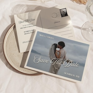 May include: A collection of wedding stationery, including a "Save The Date" postcard featuring a couple kissing by the water. The postcard includes the names Nikki & Cameron, the date October 19, 2030, and the location Houston, Texas.