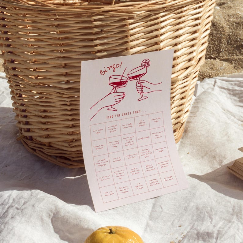 May include: A red and white bingo game card with the word "bingo" at the top. The card features illustrations of cocktails and hands, with various prompts for guests to find. A woven basket is in the background.