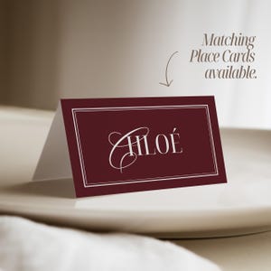 Puede incluir: Una tarjeta de mesa burdeos con el nombre "CHLO&Eacute;" en elegante escritura blanca. La tarjeta tiene un borde blanco y se muestra en un plato blanco. El texto encima de la tarjeta dice "Tarjetas de Mesa a juego disponibles."