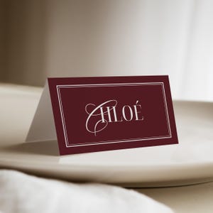 Peut inclure: Une carte de place de couleur bordeaux avec le nom "CHLOÉ" en écriture blanche élégante. La carte est bordée d'un cadre blanc et est présentée sur une assiette blanche, pour un événement formel ou un mariage.