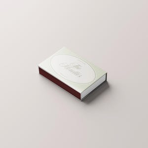 Puede incluir: Una caja de cerillas rectangular con un dise&ntilde;o verde claro y blanco. La parte superior presenta un &oacute;valo con la inscripci&oacute;n "The Morettis" en escritura elegante. La superficie de fricci&oacute;n es de color marr&oacute;n rojizo oscuro.