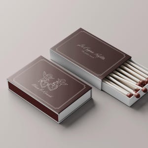 Può includere: Due scatole di fiammiferi marroni con finiture argentate e testo. Una scatola è aperta, rivelando fiammiferi bianchi con punte marroni. La scatola chiusa presenta un'illustrazione di due cherubini e cuori. Il testo sulle scatole recita "La Coppia Perfetta" e "Alessia & Riccardo."