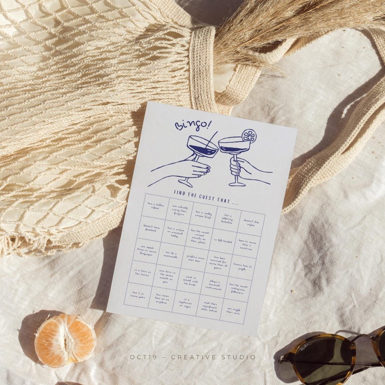 May include: A white paper game card with blue illustrations and text, including the word "Bingo!" and "Find the guest that...". The card is next to a woven bag, dried grass, and a sliced orange.