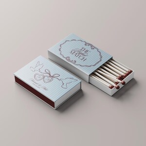 May include: Two matchboxes, one open, with a light blue and white striped design. The open box reveals matches with brown tips. The closed box features doves, hearts, and text. The text on the open box reads "The Perfect Match."
