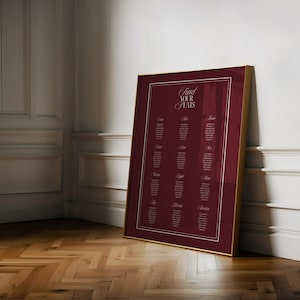 Może przedstawiać: Oprawiony bordowy plan stołów ze złotą obwódką, oparty o białą ścianę. Na wykresie widnieje napis "Find Your Seats" elegancką czcionką, z nazwiskami gości wymienionymi pod numerami od jednego do dwunastu.