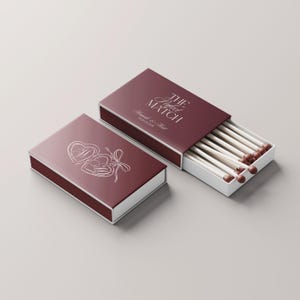 Puede incluir: Dos cajas de cerillas granate, una abierta, que muestran cerillas con puntas marrones. La caja cerrada presenta un diseño de corazón y el texto "The Perfect Match" con nombres y una fecha. Interiores blancos.