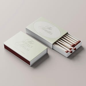 Puede incluir: Dos cajas de cerillas, una abierta, que muestran cerillas de madera con puntas rojas. Las cajas son blancas con un dise&ntilde;o a rayas verde claro y texto plateado que dice "The Perfect Match" y "The Morettis".