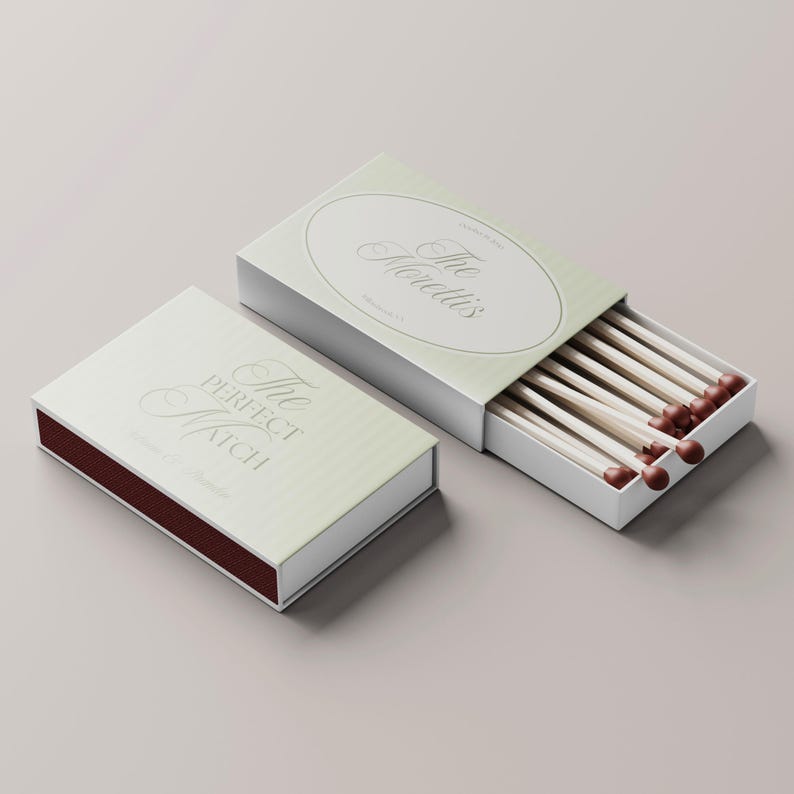 Puede incluir: Dos cajas de cerillas, una abierta, que muestran cerillas de madera con puntas rojas. Las cajas son blancas con un dise&ntilde;o a rayas verde claro y texto plateado que dice "The Perfect Match" y "The Morettis".