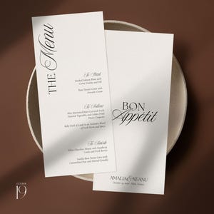 May include: Two vertical white menu cards with black serif and script typography. The left card is titled THE MENU and lists courses including Smoked Salmon Blinis and Miso Marinated Black Cod. The right card reads BON APPETIT and AMALIA & KEANU.