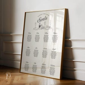 May include: A vertical rectangular wedding seating chart in a thin gold frame. The white sign features the text "Please find your seats" above a line drawing of a table setting. It lists twelve tables with guest names in black serif font.