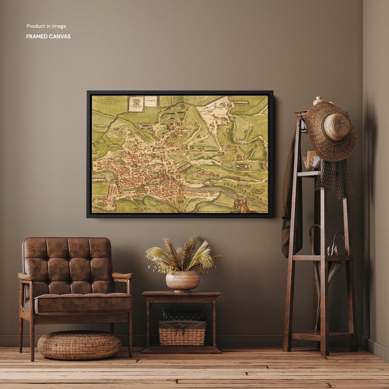 Op de afbeelding: Ingelijste canvas kunst met een vintage kaart in een zwart frame. De kaart toont een gedetailleerde illustratie van een stad met rode gebouwen en groene landschappen. Het kunstwerk hangt aan een bruine muur, samen met een bruine stoel en een houten kapstok.