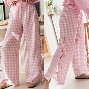 K&ouml;nnte beinhalten: Weite Hose mit rosa-wei&szlig;em Vichy-Karo-Muster. Die Hose hat einen Kordelzug in der Taille und Seitenschlitze mit rosa Schleifen. Der Stoff scheint eine leichte Baumwoll- oder Leinenmischung zu sein.