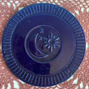 May include: A circular, deep blue ceramic coaster featuring a raised relief design in the center. The design depicts a crescent moon, a sun with radiating rays, and two small stars. The outer rim has a textured, grooved pattern around the edge.