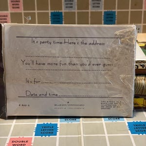 May include: A white invitation card with text that reads "It's party time - Here's the address" and "You'll have more fun than you'd ever guess." The card is wrapped in clear plastic and sits on a Scrabble board.