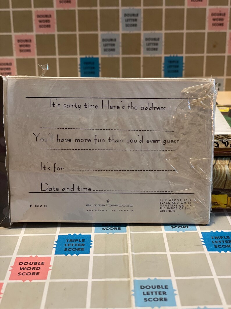 May include: A white invitation card with text that reads "It's party time - Here's the address" and "You'll have more fun than you'd ever guess." The card is wrapped in clear plastic and sits on a Scrabble board.