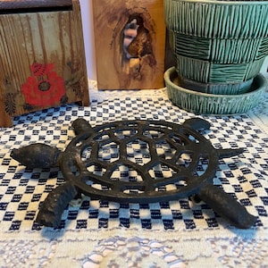May include: A cast iron turtle-shaped trivet with a honeycomb design. The trivet is black and sits on a blue and white checkered tablecloth. Other items in the background include a wooden box with a red flower design and a green ceramic planter.