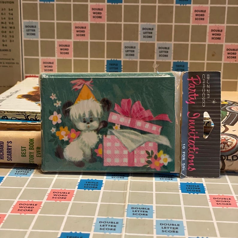 May include: A stack of party invitations with a cartoon puppy wearing a party hat and a pink gift box on the front. The invitations are packaged in a clear plastic wrap. A vintage Scrabble board is in the background.