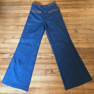 May include: High-waisted blue denim jeans featuring a wide-leg silhouette and a raw, unfinished hem. The pants include two front pockets with brown leather trim accents and matching leather detailing along the waistband.