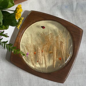 May include: Square wooden coaster with a clear resin top. The resin contains wheat stalks, small orange flowers, and a red ladybug. The coaster has a gold-colored background and is surrounded by a dark brown wooden frame.
