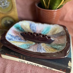 May include: A square ceramic ashtray with rounded corners, featuring a glossy dark brown rim and a central dark brown cigar rest. The interior surface displays a vibrant, multi-colored drip glaze pattern in shades of turquoise, yellow, and lavender.