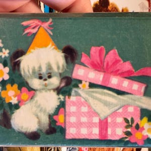 May include: A vintage birthday card featuring a cartoon puppy wearing a party hat. The puppy is next to a pink gift box with a bow. The background is teal with flowers.