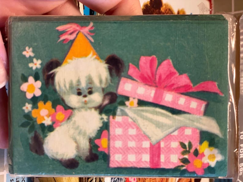 May include: A vintage birthday card featuring a cartoon puppy wearing a party hat. The puppy is next to a pink gift box with a bow. The background is teal with flowers.