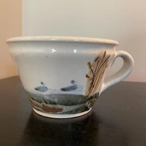 May include: A white ceramic mug with a brown handle and a nature scene painted on the side. The scene features a brown tree with green leaves and three blue birds in flight.