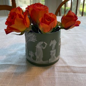 May include: A small green ceramic vase with a white design of people and trees, filled with orange roses.