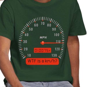 May include: A dark green t-shirt featuring a graphic of a speedometer with white markings, a red needle pointing past 110 MPH, a digital odometer reading 010075, and a red rectangular box containing the text "WTF is a km/h?" printed in black.