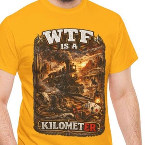 Può includere: T-shirt giallo senape con un design grafico raffigurante un treno a vapore, un paesaggio desertico e la scritta "WTF IS A KILOMETER". Il design include un saloon, carte da gioco e stivali da cowboy.