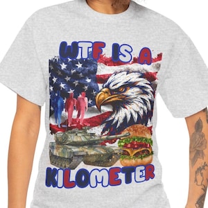 Può includere: T-shirt grigio chiaro con un design grafico. Il design include una bandiera americana, un'aquila, un carro armato, un hamburger e il testo "WTF IS A KILOMETER" in blu e rosso. La maglietta ha un girocollo.