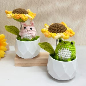 May include: Two crochet plant decorations. One features a pink bunny with a sunflower, the other a green frog with a sunflower. Both are in white geometric pots with green faux grass.