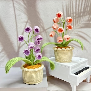 May include: Two crochet flower arrangements in beige pots. One has purple bell-shaped flowers with green stems and leaves. The other has peach-coloured flowers. Both are on a white surface, with a miniature piano in the background.