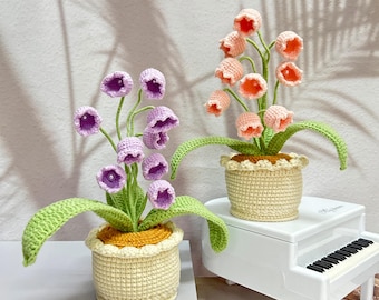 May include: Two crochet flower arrangements in beige pots. One features purple bell-shaped flowers with green stems and leaves. The other has peach-colored flowers. Both are on a white surface, with a miniature piano in the background.