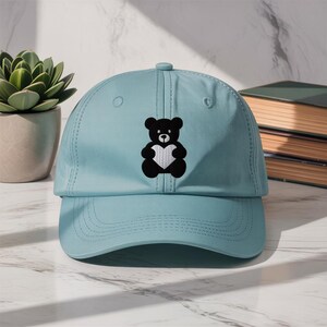 Pode incluir: Boné de beisebol azul claro com um urso de pelúcia preto segurando um coração branco bordado na frente. O boné tem uma viseira curva e um botão na parte superior. Acessório casual.