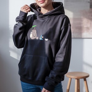 May include: Black hooded sweatshirt with a front pocket. The sweatshirt features a cartoon illustration of a gray creature with a green leaf, a blue creature, and a white creature.