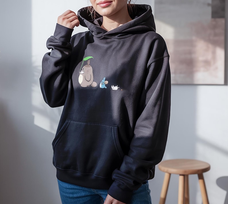 May include: Black hooded sweatshirt with a front pocket. The sweatshirt features a cartoon illustration of a gray creature with a green leaf, a blue creature, and a white creature.