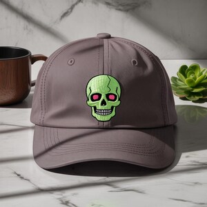 May include: A grey baseball cap with an embroidered neon green skull and pink eyes. The skull design is on the front of the cap. The cap has a curved peak and a button on top.